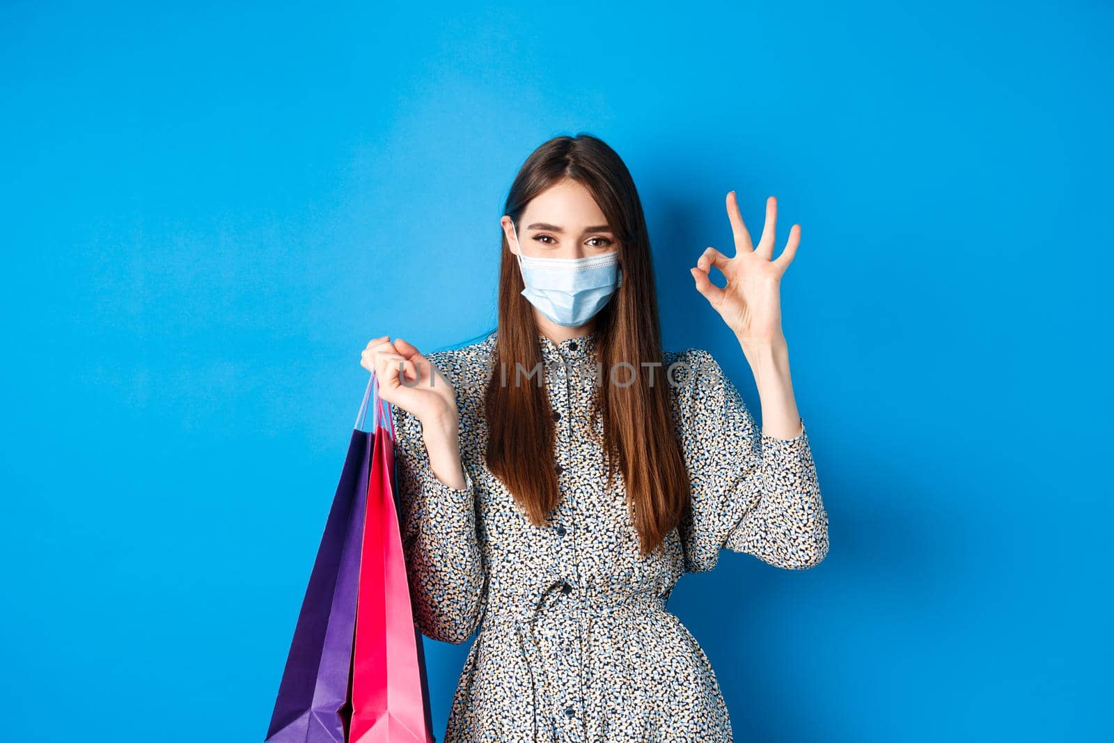 Covid-19, pandemic and lifestyle concept. Attractive woman wear medical mask on shopping, show okay and hold bags with purchases, blue background.