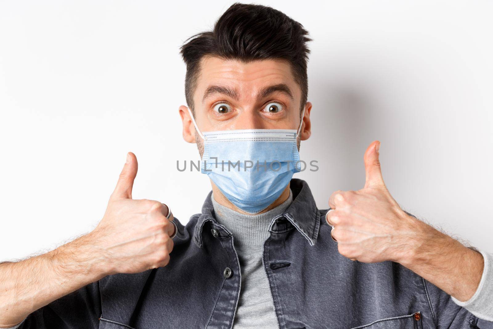 Covid-19, health and lifestyle concept. Close-up of excited positive guy in medical mask supports wearing medical mask during pandemic, showing thumbs up in approval.