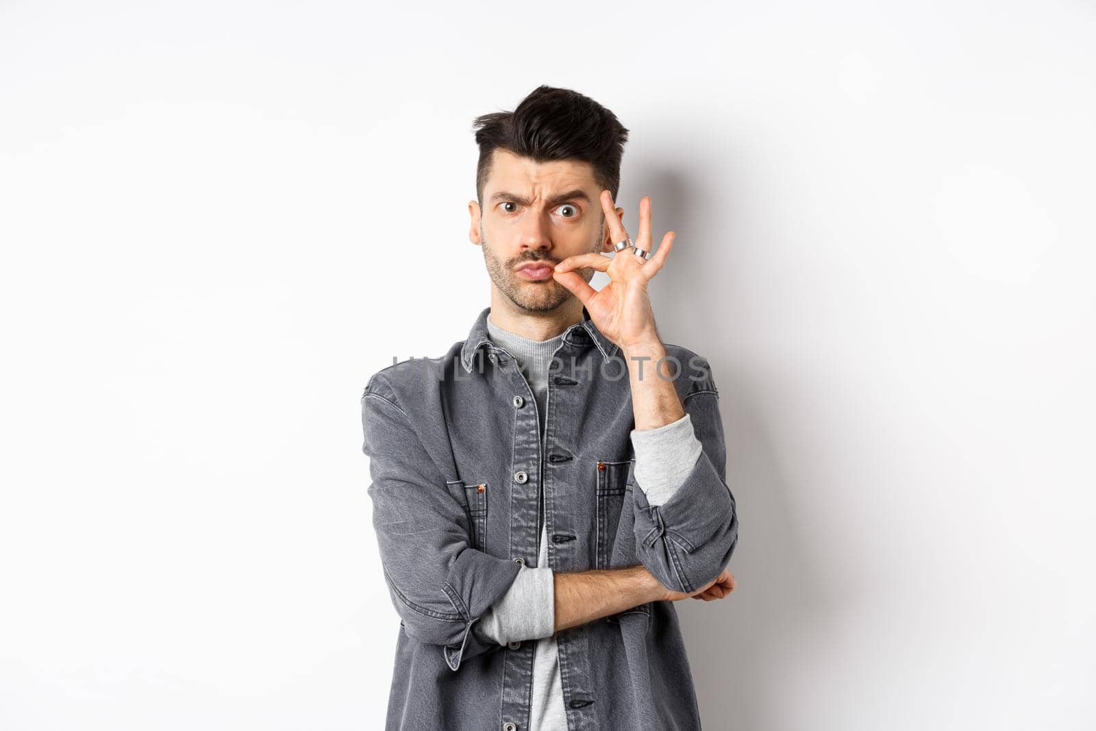 Serious man making zip gesture, tell to keep mouth shut, frowning and make seal on lips, standing on white background.