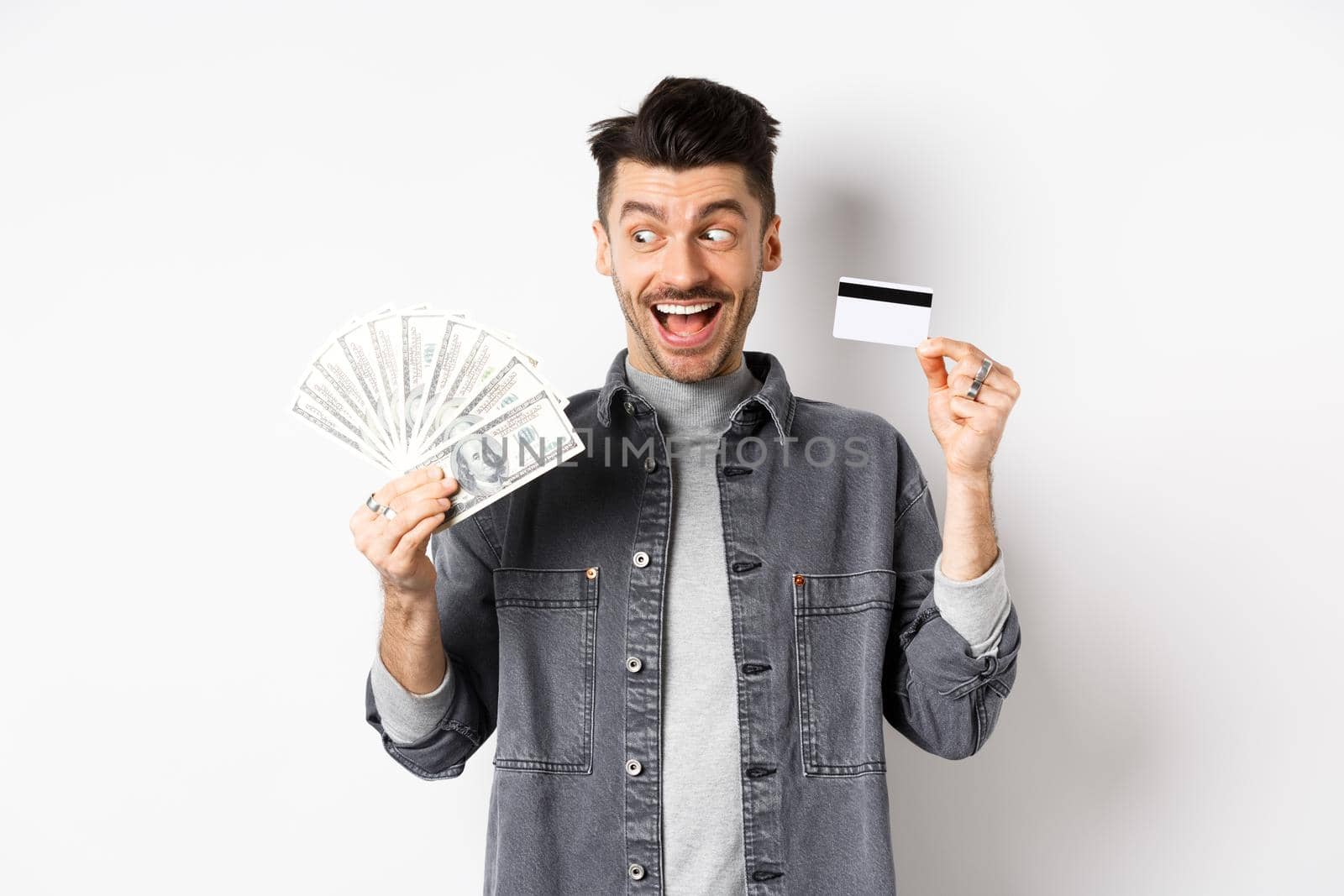 Excited guy holding plastic credit card and dollar bills, standing amused on white background.