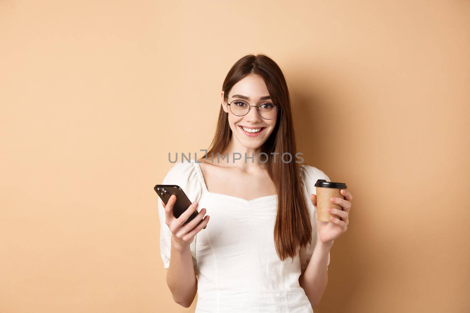 Happy caucasian woman in glasses standing with cup of coffee and smatphone, standing on beige background. Copy space