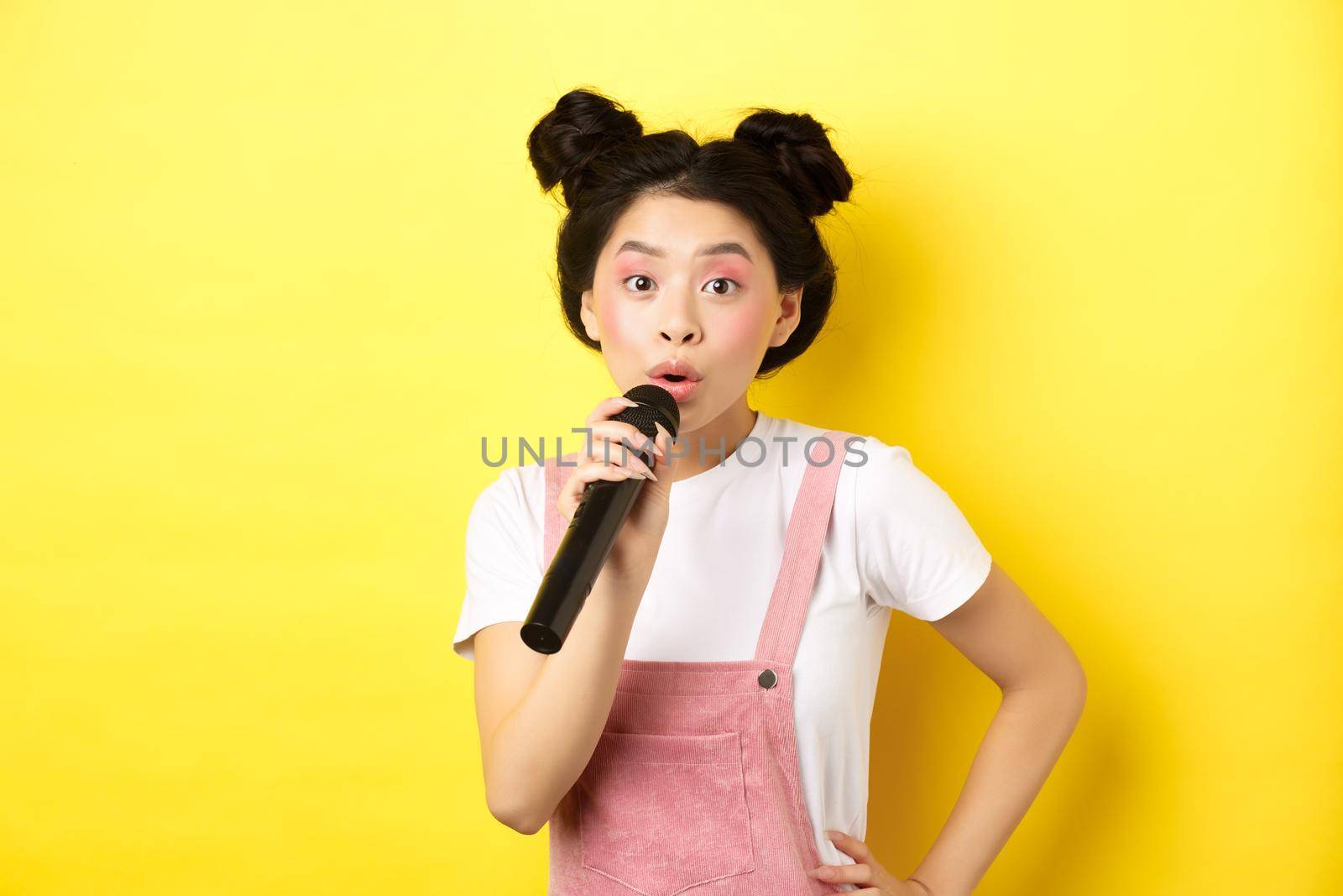 Cute asian teen girl with bright makeup, singing in microphone karaoke, standing against yellow background.