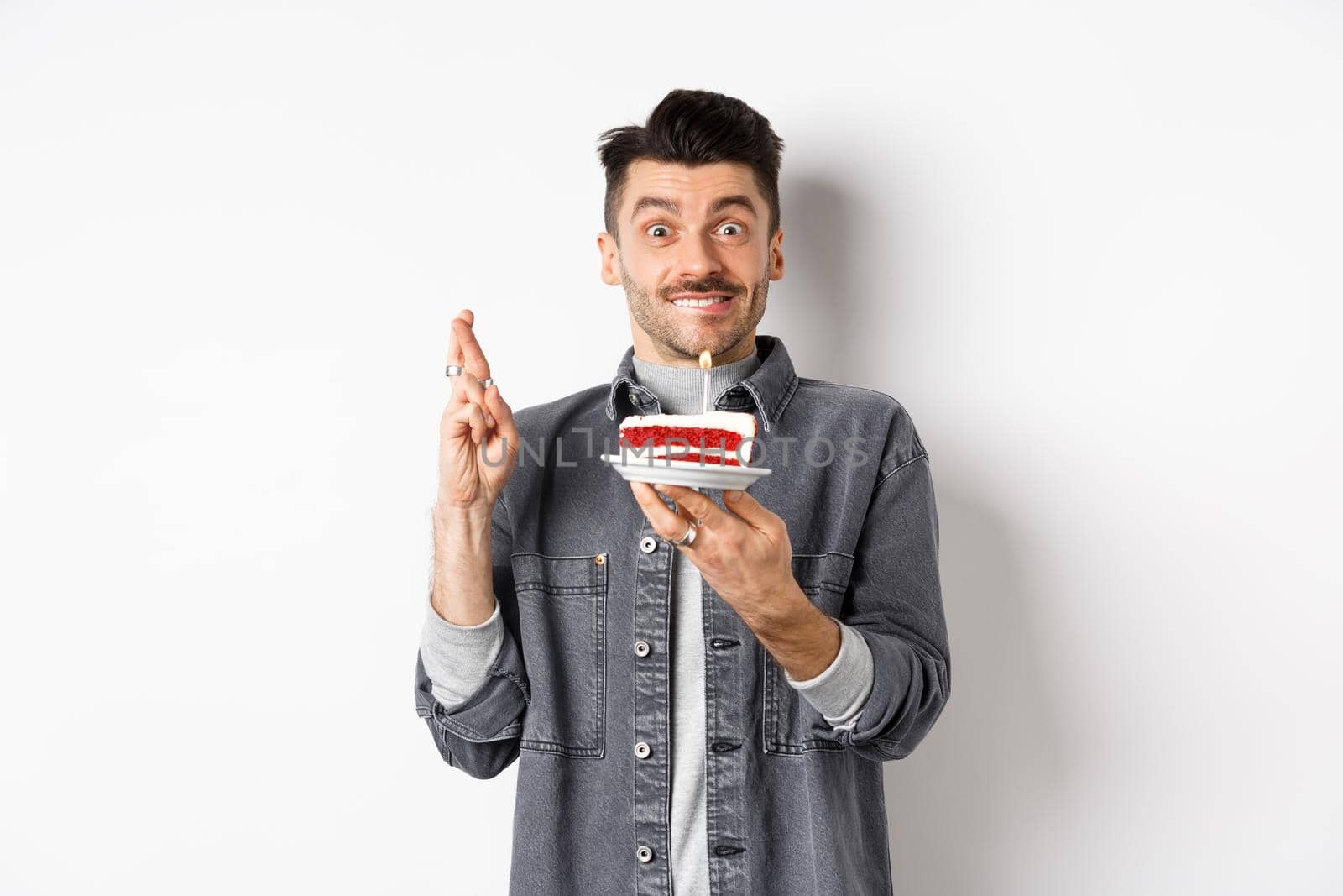 Birthday. Happy guy making wish with fingers crossed, holding bday cake with candle, standing on white background.