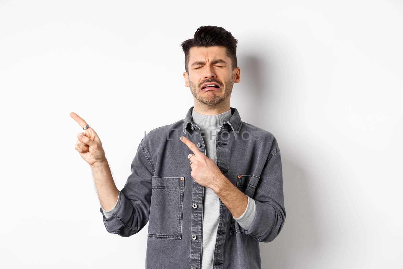 Sad crying man pointing fingers left at logo, sobbing and whining, being jealous or upset, standing on white background.