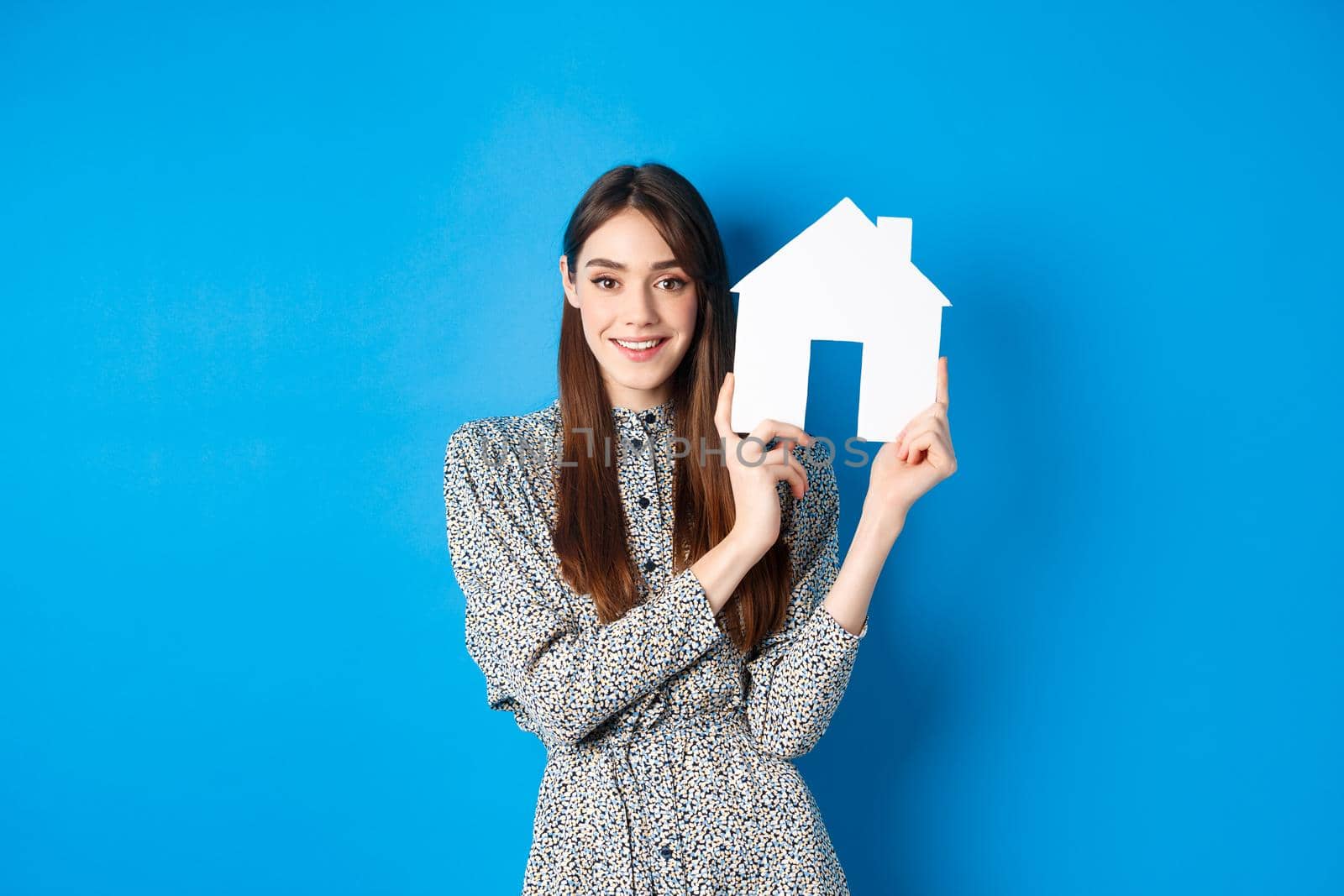 Real estate, realtors and insurance concept. Beautiful lady in dress showing paper house cutout, suggest property for sale, standing on blue background.