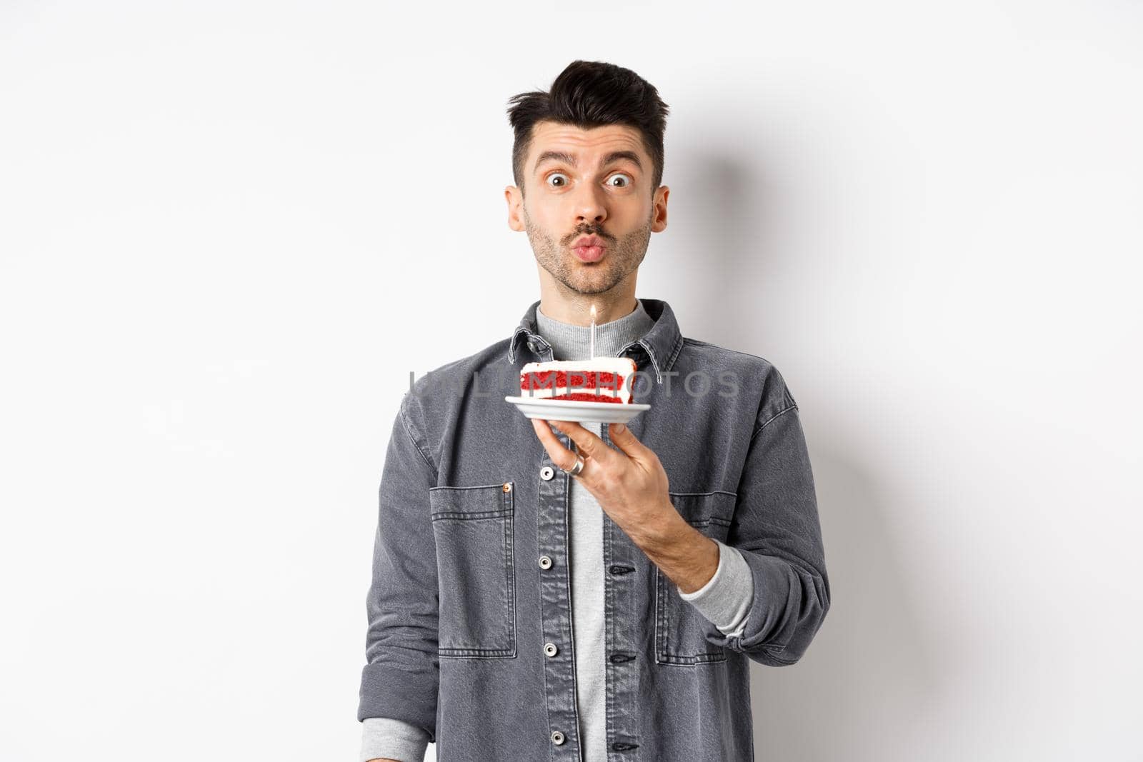 Man celebrating birthday, blowing candle on cake and making wish, standing on white background.