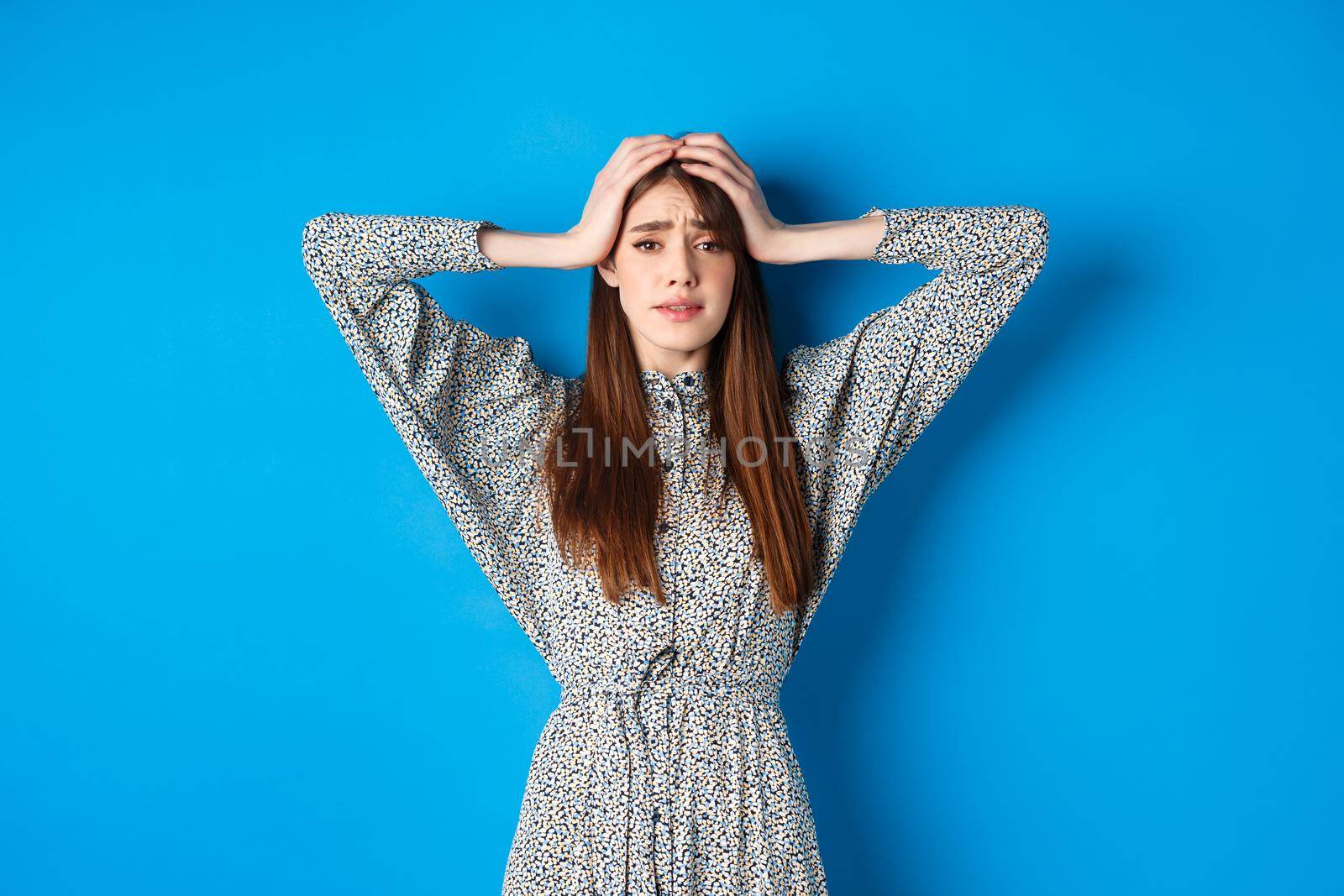 Frustrated adult woman with natural hair holding hands on head and panicking, looking alarmed and scared, standing in dress against blue background.