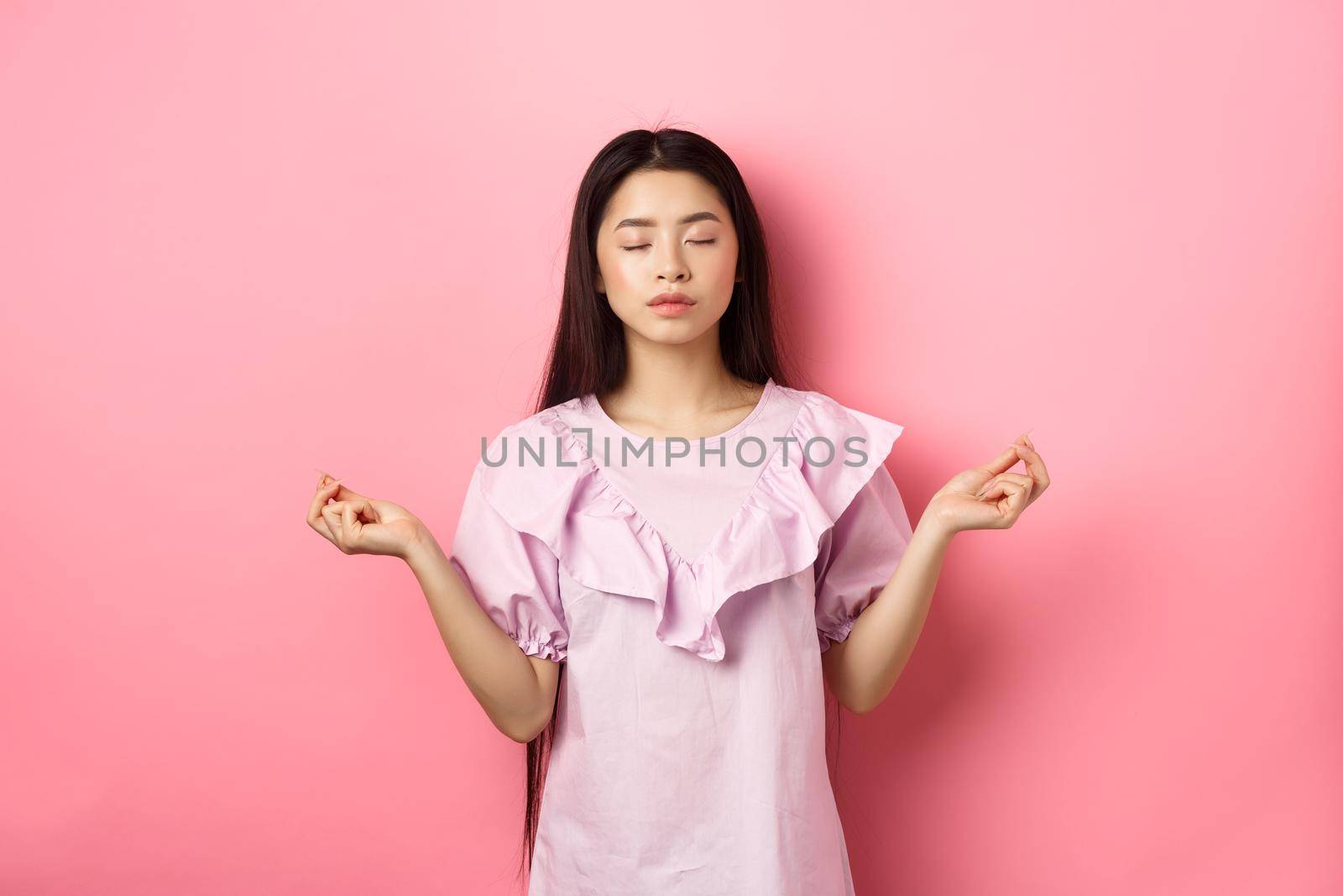 Stay calm. Peaceful asian woman meditating, hold hans in zen gseture and close eyes, practice yoga, standing patient on pink background.