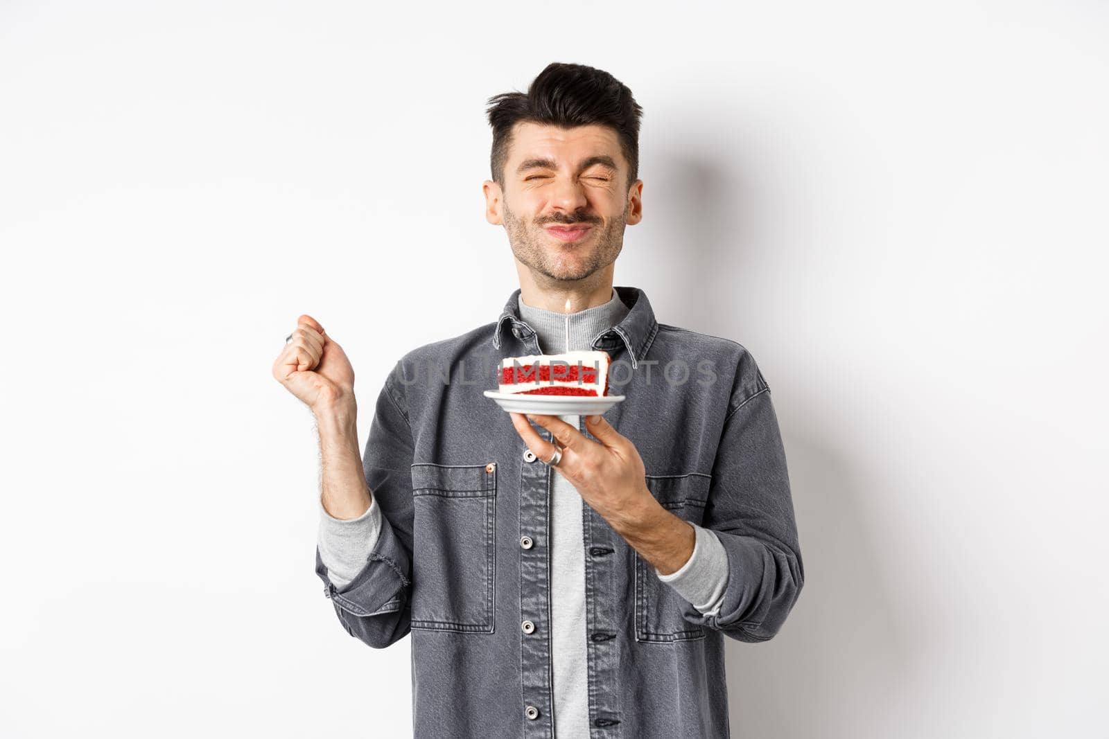 Happy birthday guy making wish on cake with candle, celebrating bday, standing on white background.