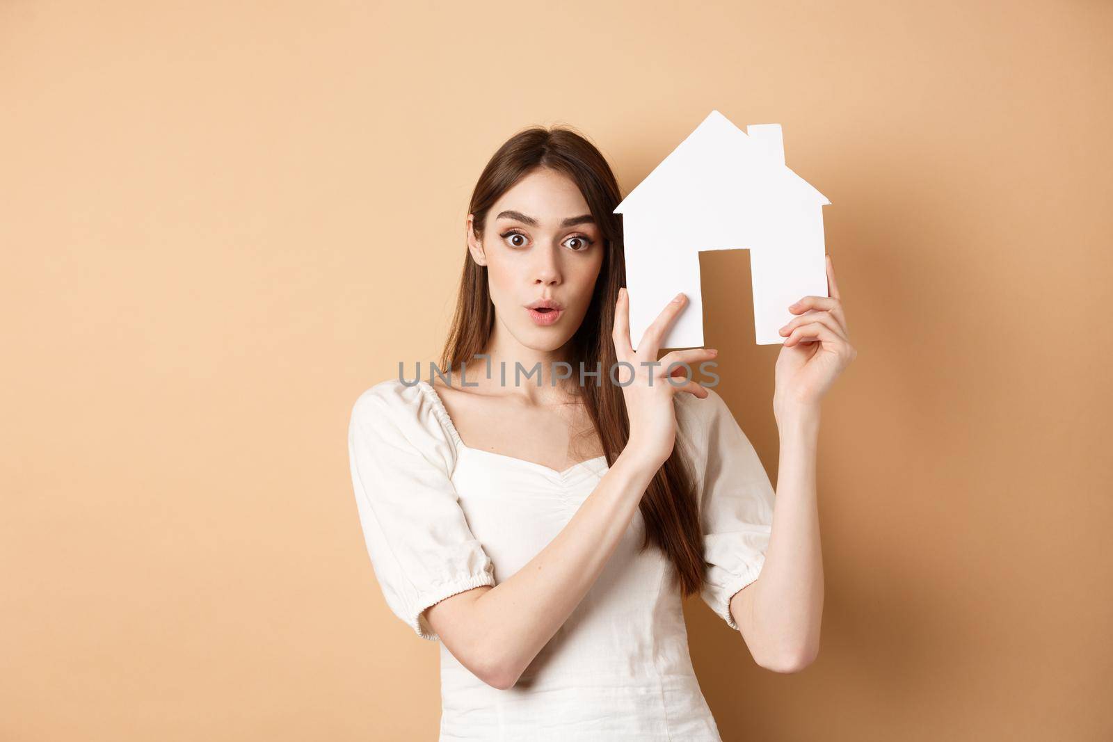 Real estate. Excited young woman showing house cutout and looking at camera, renting property, standing on beige background.