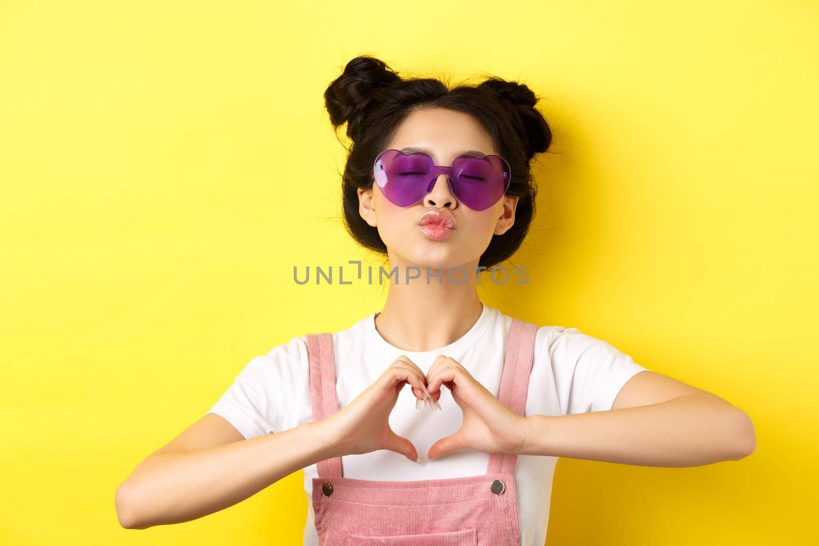 Summer and fashion concept. Cute glam girl with sunglasses and hairbuns, showing heart sign and pucker lips for kiss, I love you gesture, yellow background.