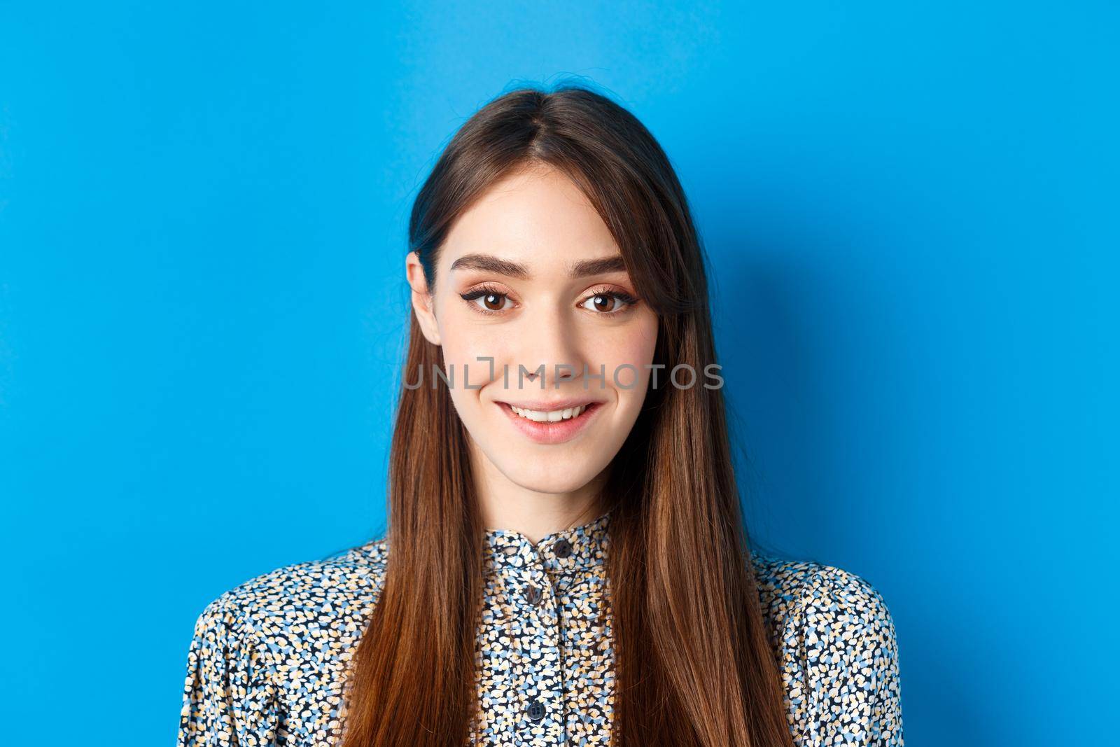 Close-up portrait of attractive natural girl with long hair and makeup, smiling happy and friendly at camera, blue background.