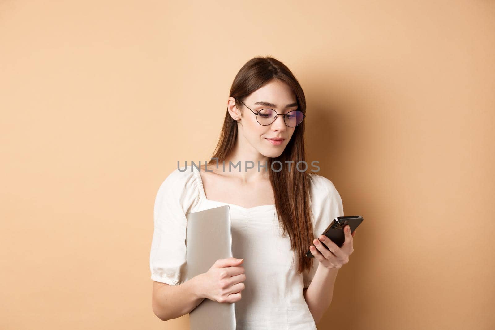 Young girl in glasses chatting on mobile phone and holding laptop, working, standing on beige background.