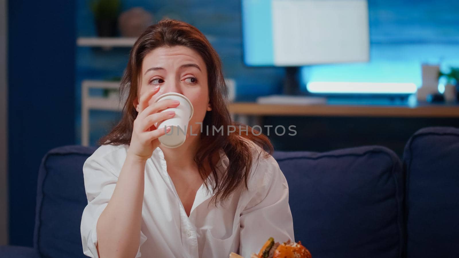 Close up of businesswoman eating fast food meal on couch with movie on TV. Hungry adult enjoying burger from takeaway order while watching television in living room. Person with free time