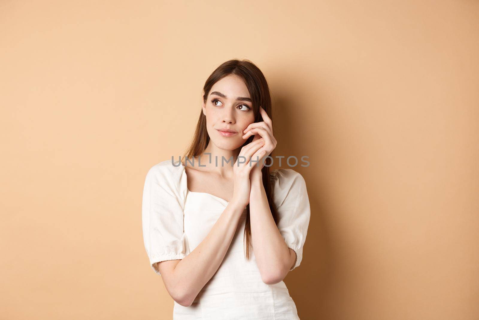 Dreamy and romantic girl looking aside with thoughtful gaze, touching clean gentle facial skin, standing against beige background.