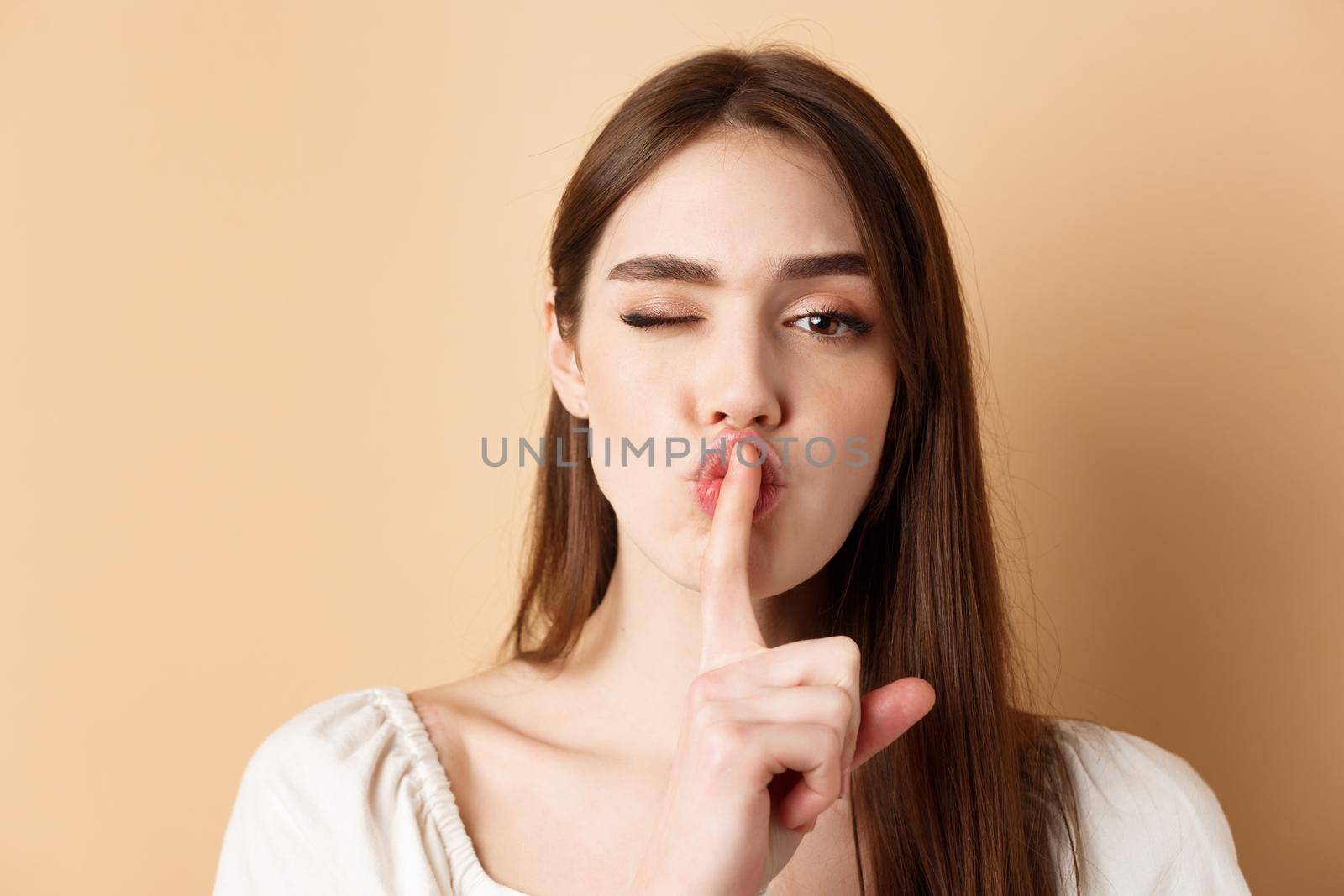 Close up of attractive woman shushing at camera, hiding secret, making hush sign and winking, show taboo sign, standing on beige background.