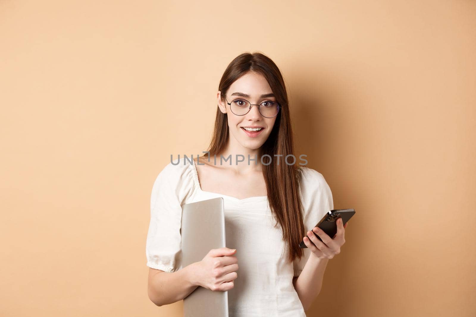 Smiling young woman in glasses reading exciting news on mobile phone, holding laptop and looking happy at camera, standing on beige background.