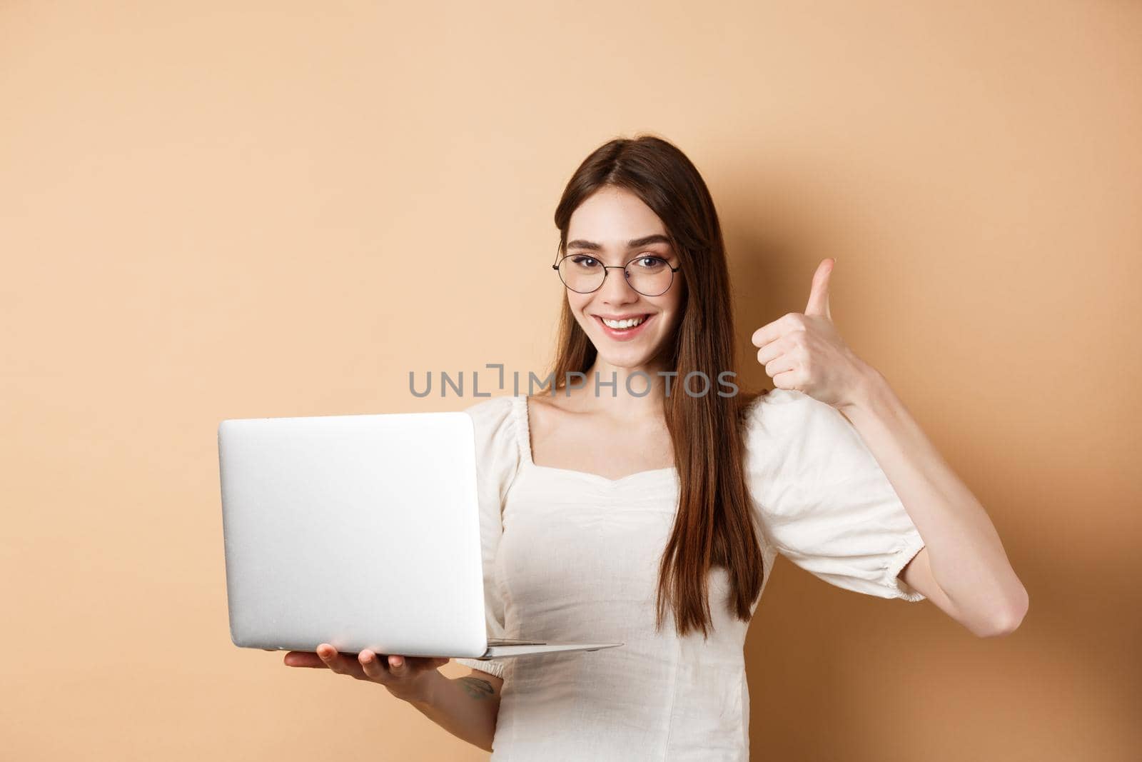 Smiling woman in glasses using laptop and showing thumb up, working on computer, standing on beige background.