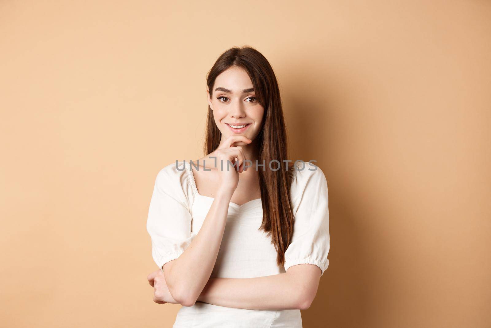 Smiling young european woman in dress, having an idea, listening with interest, touching chin and looking at camera intrigued, standing against beige background.