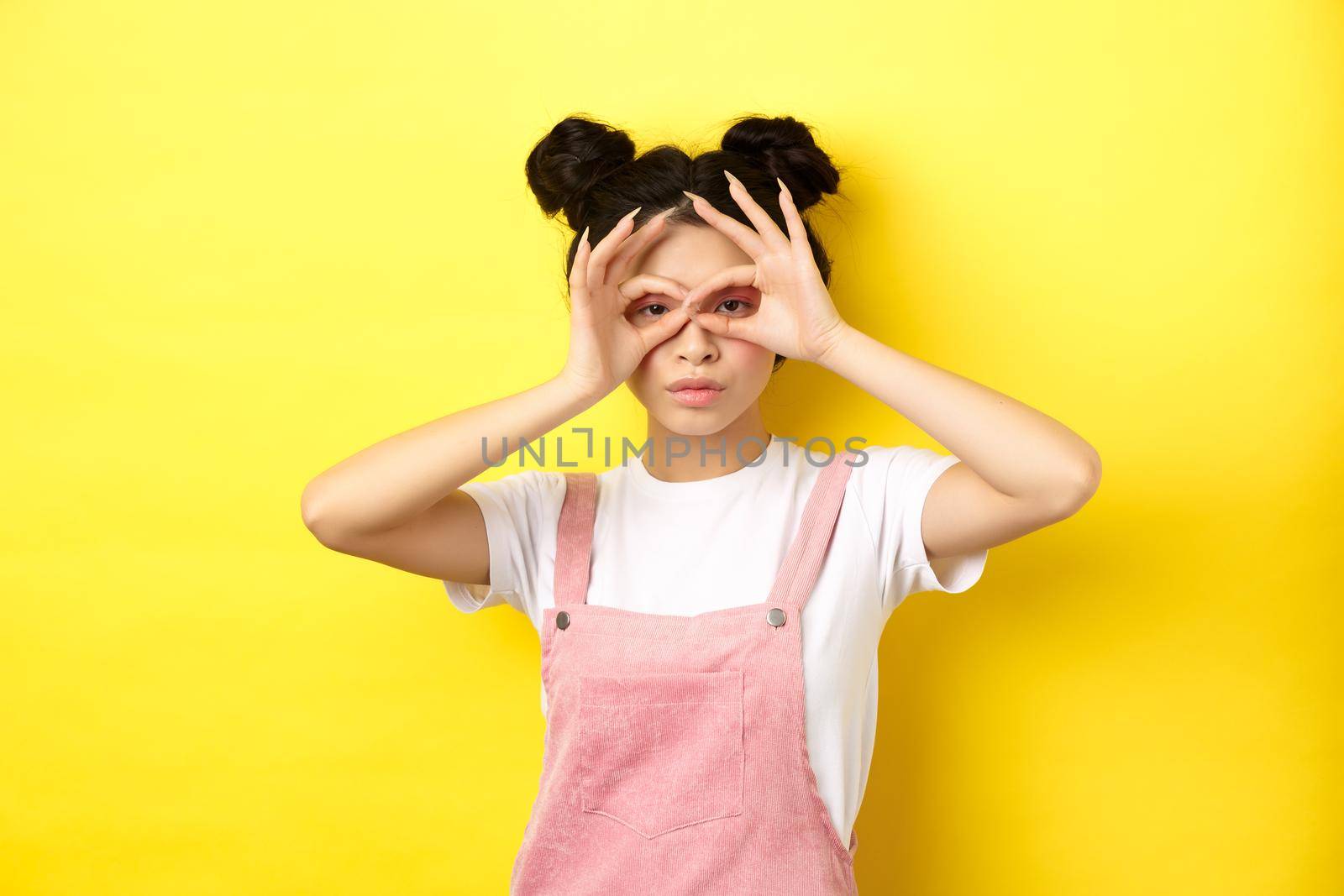 Funny asian girl look through hand binoculars, watching you, standing in summer clothes on yellow background.