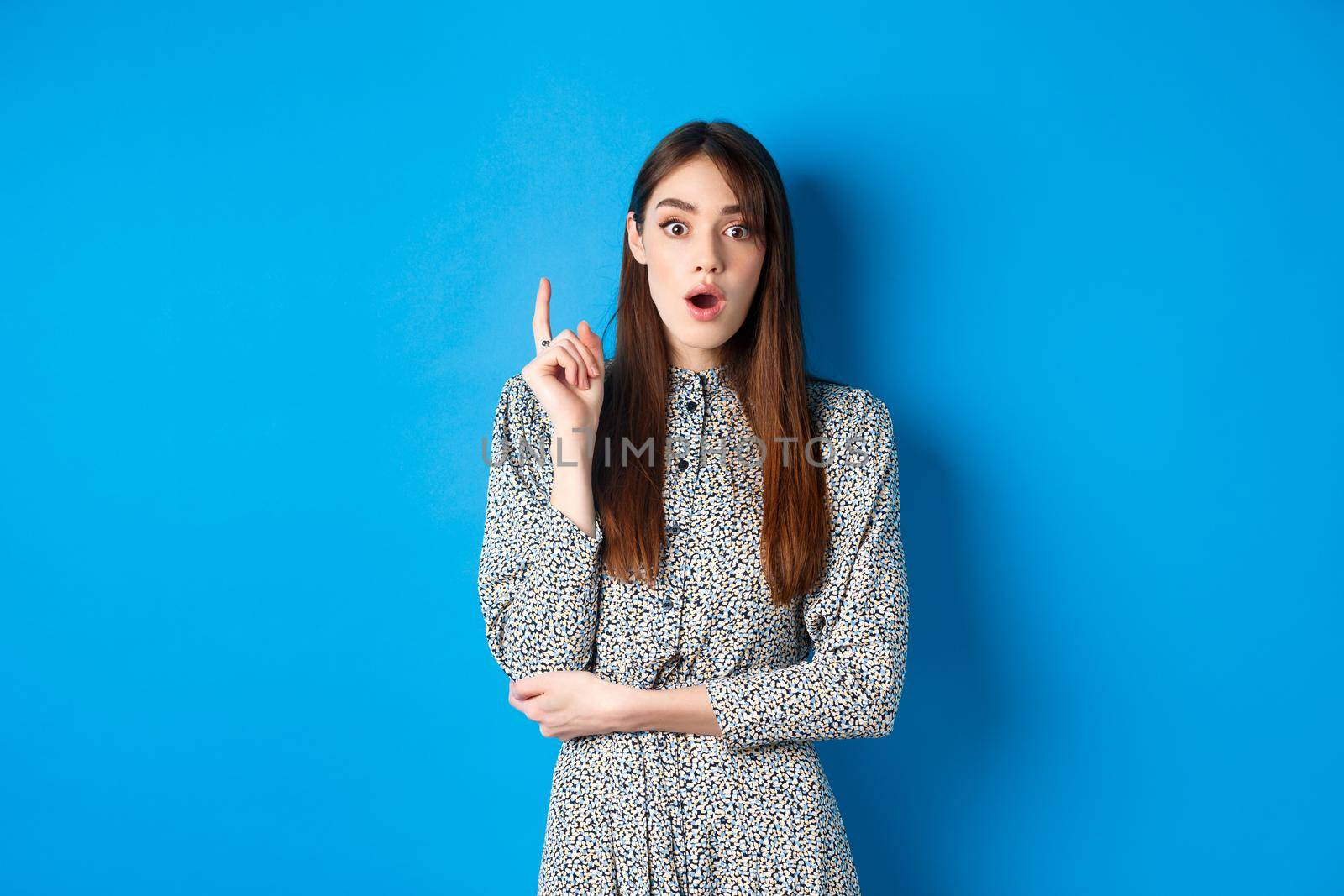Excited female model in dress raising finger, pitching idea and look amazed at camera, found solution or have plan, understood something, standing on blue background.
