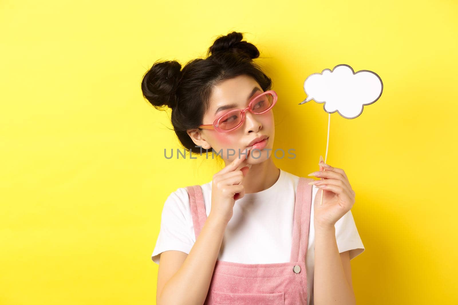 Summer and fashion concept. Stylish glamour asian girl in sunglasses, holding comment cloud party mask and looking aside, yellow background.