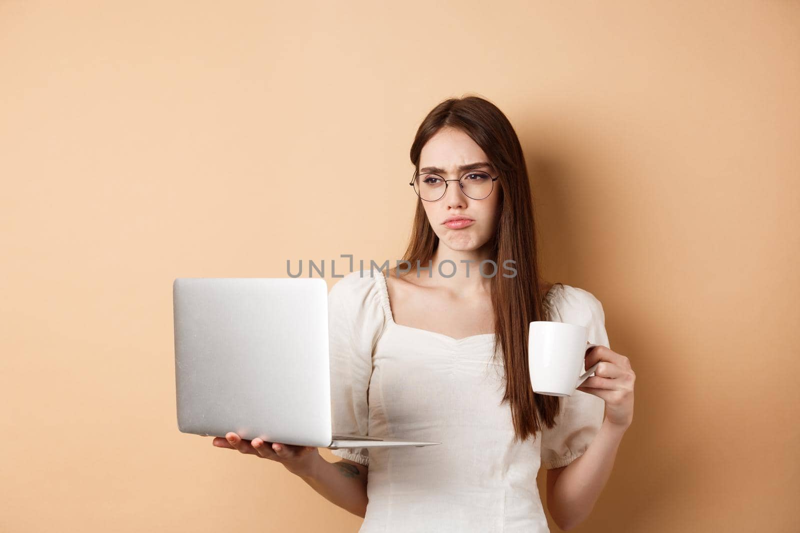 Tired working girl looking at laptop bored, drinking coffee while using computer for work, standing on beige background.
