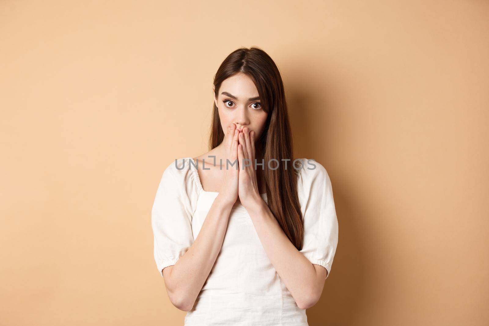 Omg shocking. Speechless woman covering mouth with hands and staring shocked at camera, gossiping, hear stunning news, beige background.