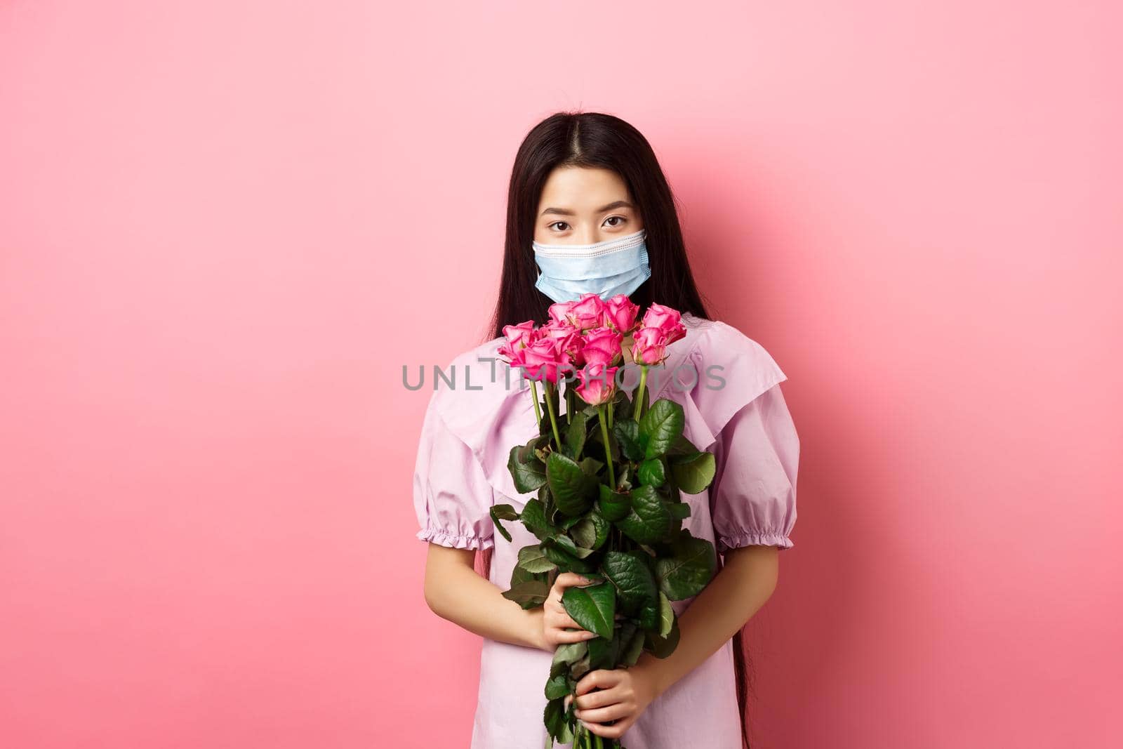 Young asian girl in medical mask holding flowers on Valentines day, receive bouquet of roses from lover, standing on pink background. Social distancing and covid-19 concept.