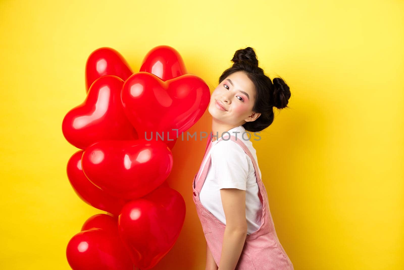 Valentines day concept. Stylish teenage asian girl posing near red hearts balloons, wear outfit for romantic date, standing happy on yellow background.