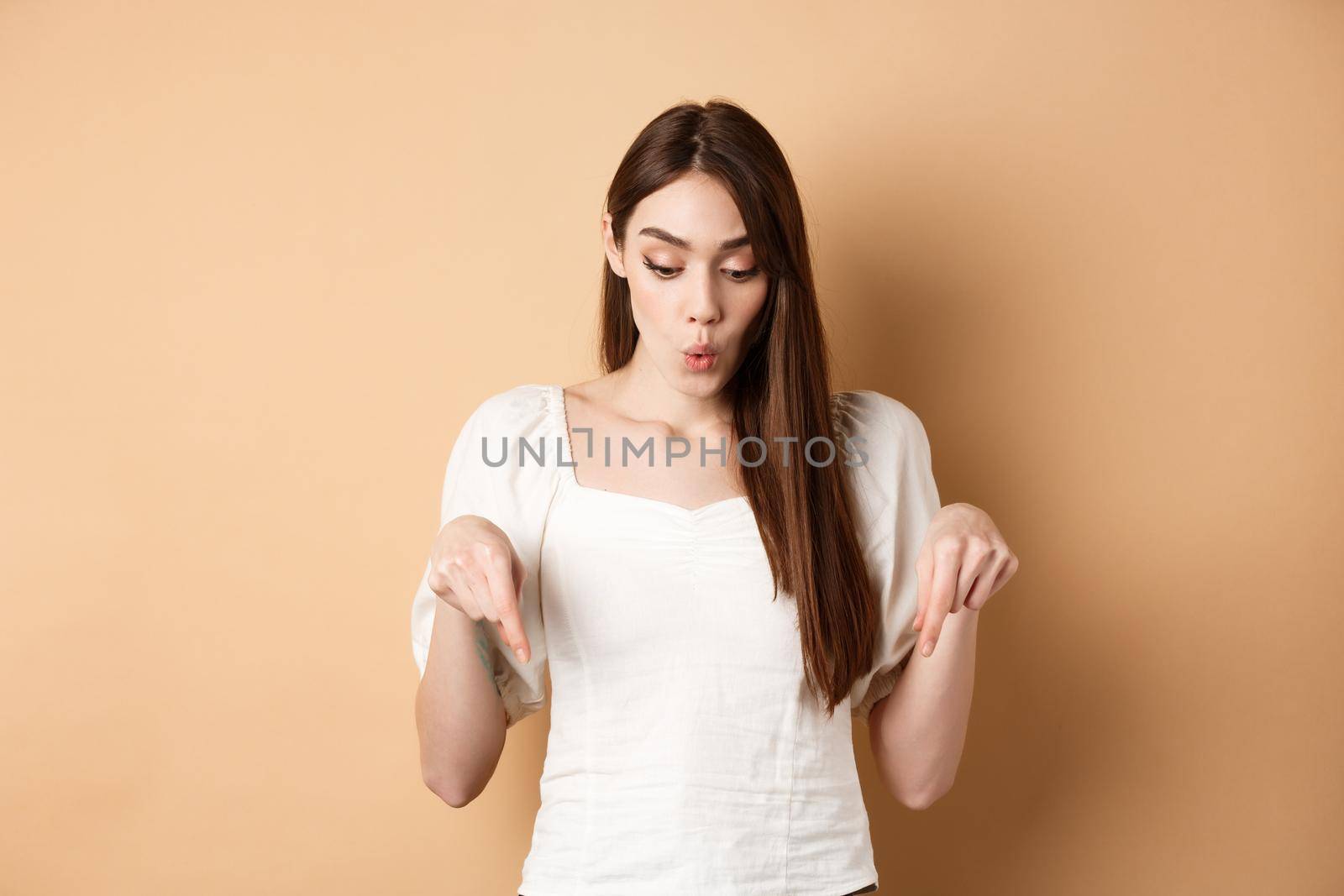 Wow look. Amazed beautiful girl checking out promo deal, pointing fingers down and gasping astonished, showing advertisement, beige background.