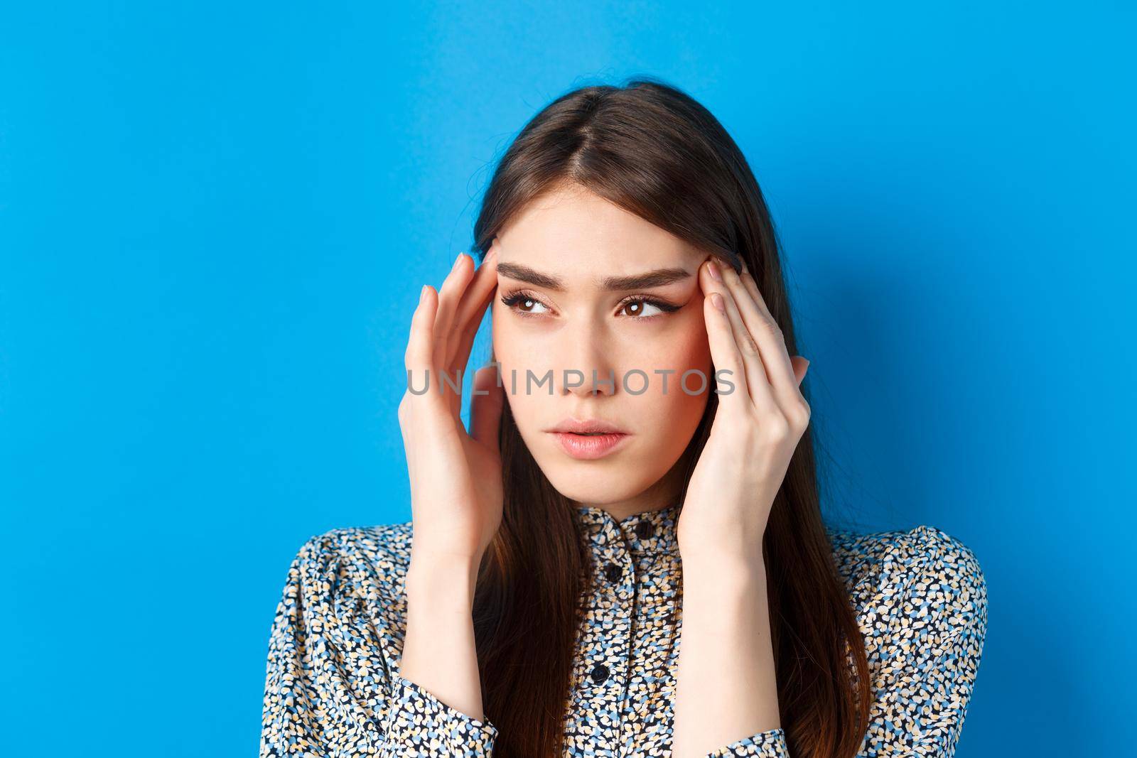 Close-up portrait of young woman feel sick, touching head temples and frowning from headache, having migraine, looking aside at logo, blue background.
