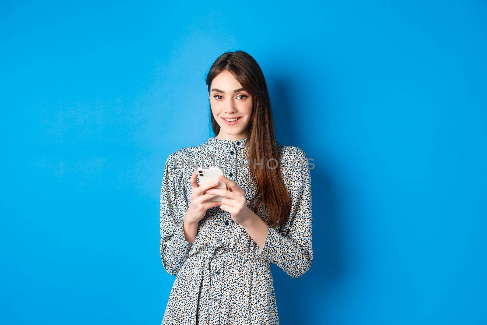 Attractive caucasian woman in dress using mobile phone and smiling, standing in dress against blue background.