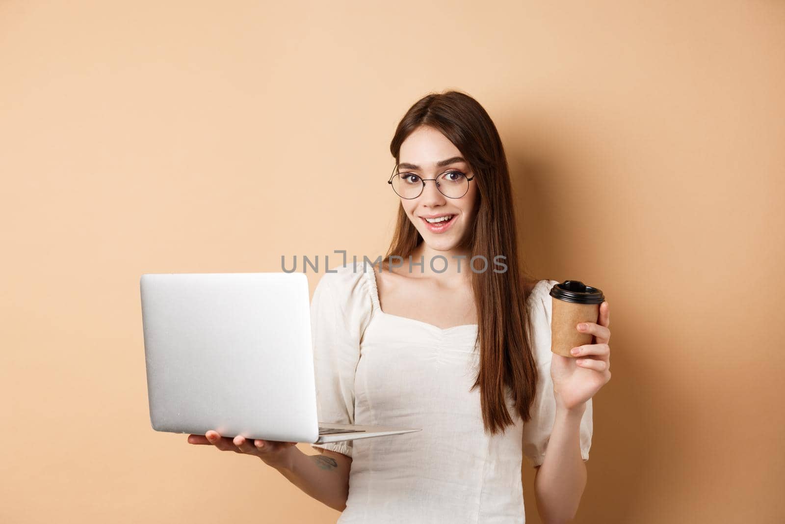 Smiling girl in glasses working on laptop and drinking coffee takeaway. Freelancer using computer at work, standing on beige background.