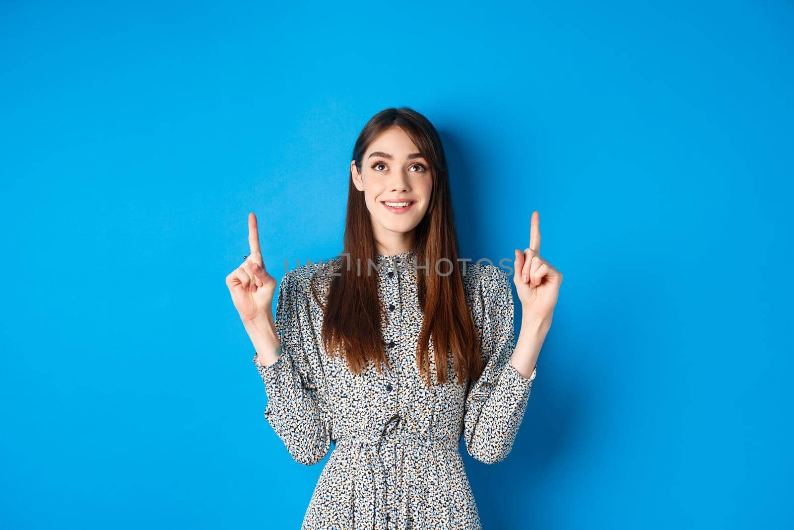 Beautiful and tender young woman pointing fingers up, smiling while advertising promo deal on blue background.