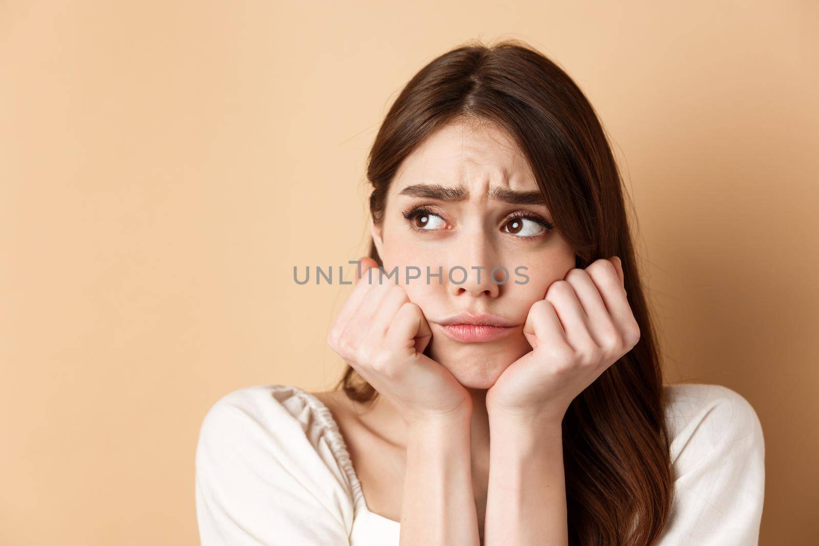 Close up of sad and gloomy girl looking aside, sulking and frowning upset, standing miserable on beige background.