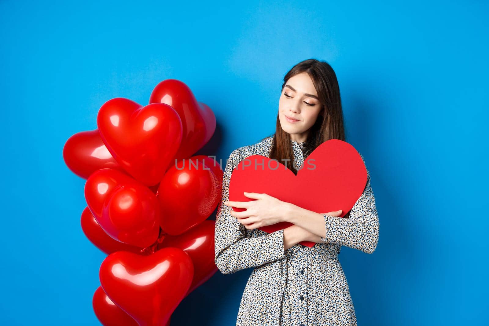 Valentines day. Romantic pretty woman in dress hugging big red heart cutout and looking dreamy, thinking of love, standing on blue background.
