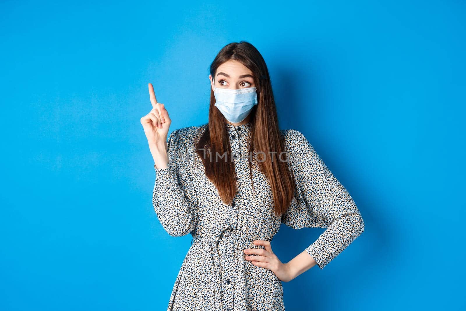 People, covid and quarantine concept. Excited cute female in medical mask, pointing and looking at logo with happy amazed face, standing on blue background.
