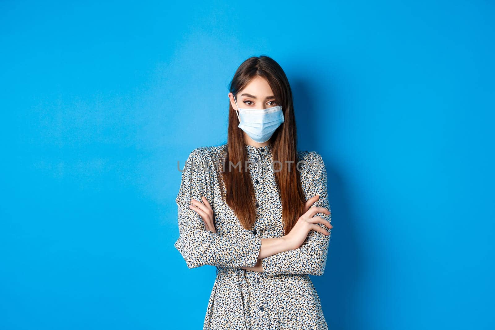 Covid-19, pandemic lifestyle concept. Serious young woman with long natural hair, wearing medical mask, cross arms on chest, protect herself from virus, blue background.