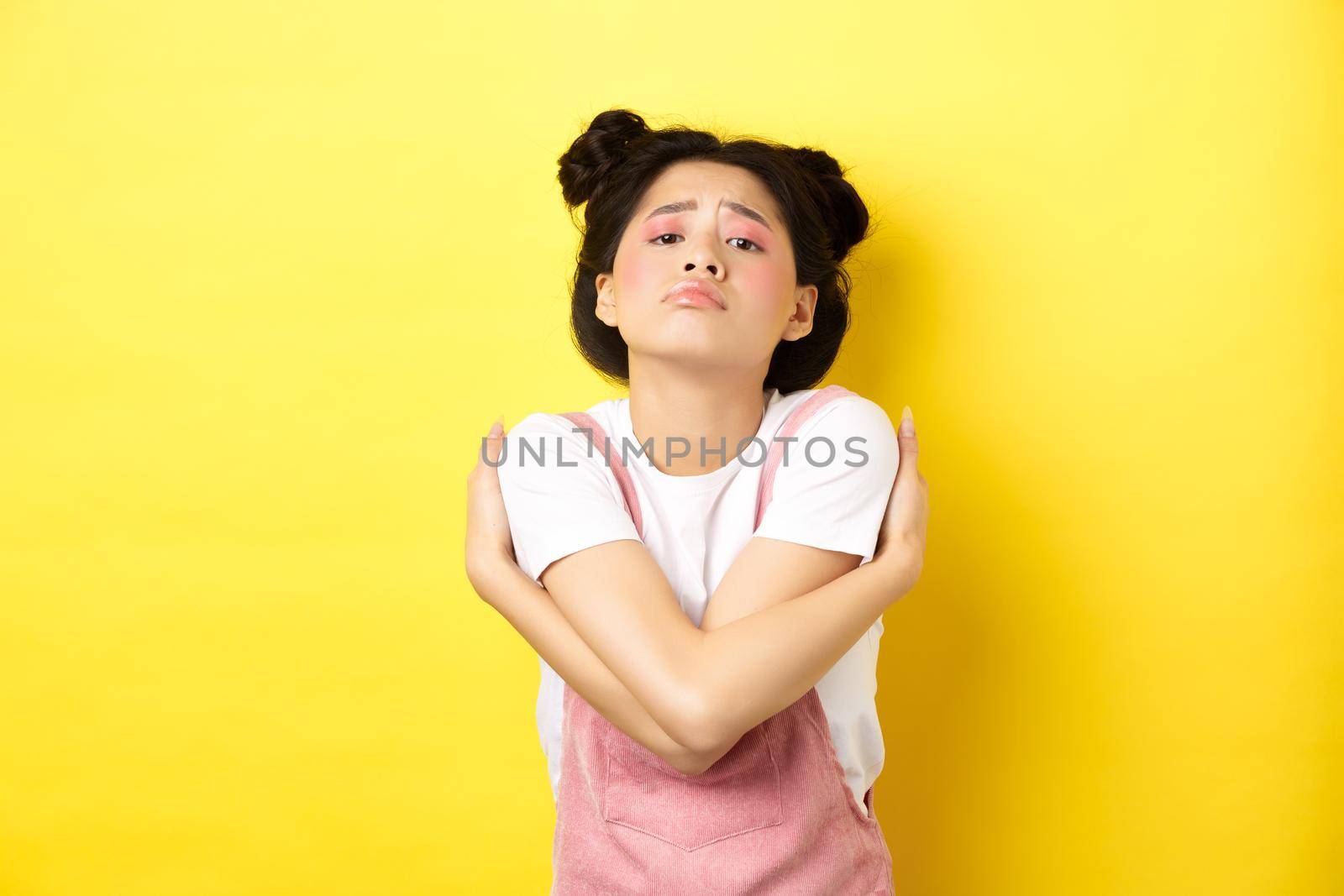 Lonely sad teen girl hugging herself, wanting to relationship and cuddles, standing alone on yellow background.
