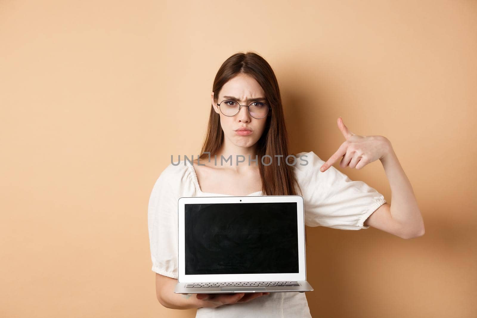 Angry frowning woman in glasses pointing at laptop screen, complaining and look disappointed, standing on beige background.