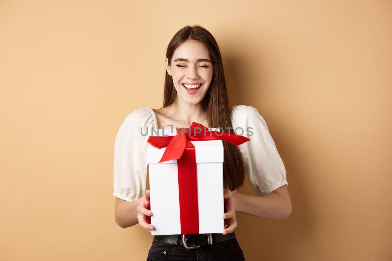 Happy smiling woman laughing, holding gift box from lover, enjoying romantic date on Valentines day, standing on beige background.