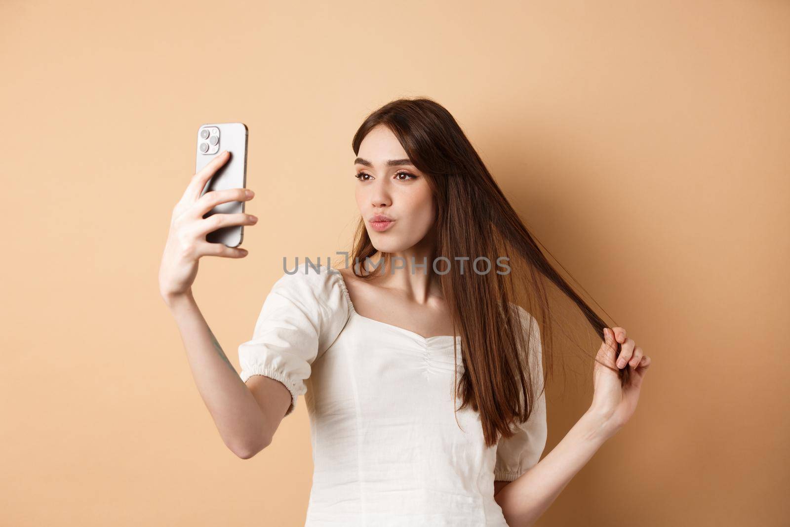 Stylish young woman playing with hair and taking selfie on smartphone, make photo for social media, standing on beige background by Benzoix