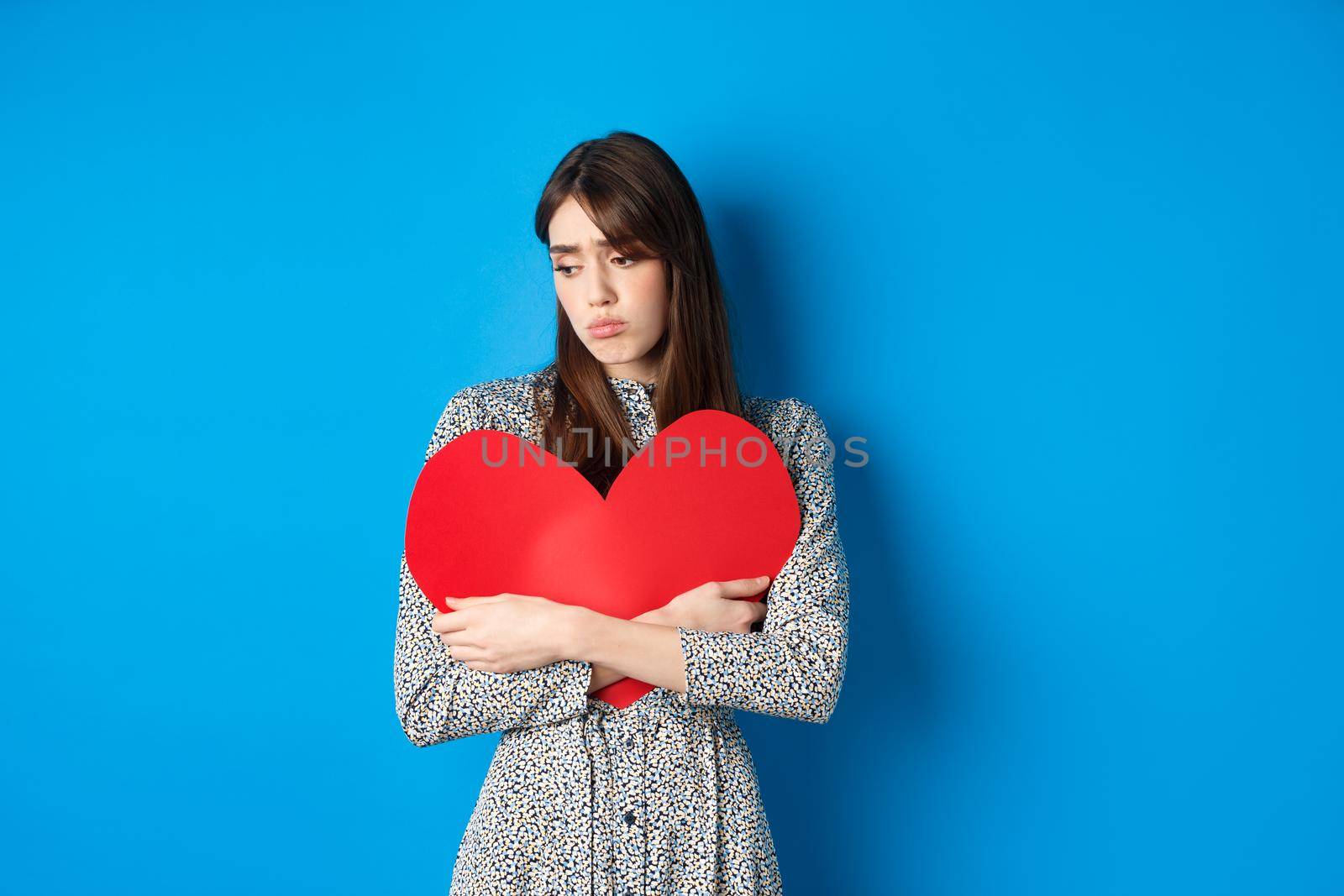 Valentines day. Lonely single girl hugging big red heart cutout and looking down upset, standing gloomy on blue background.