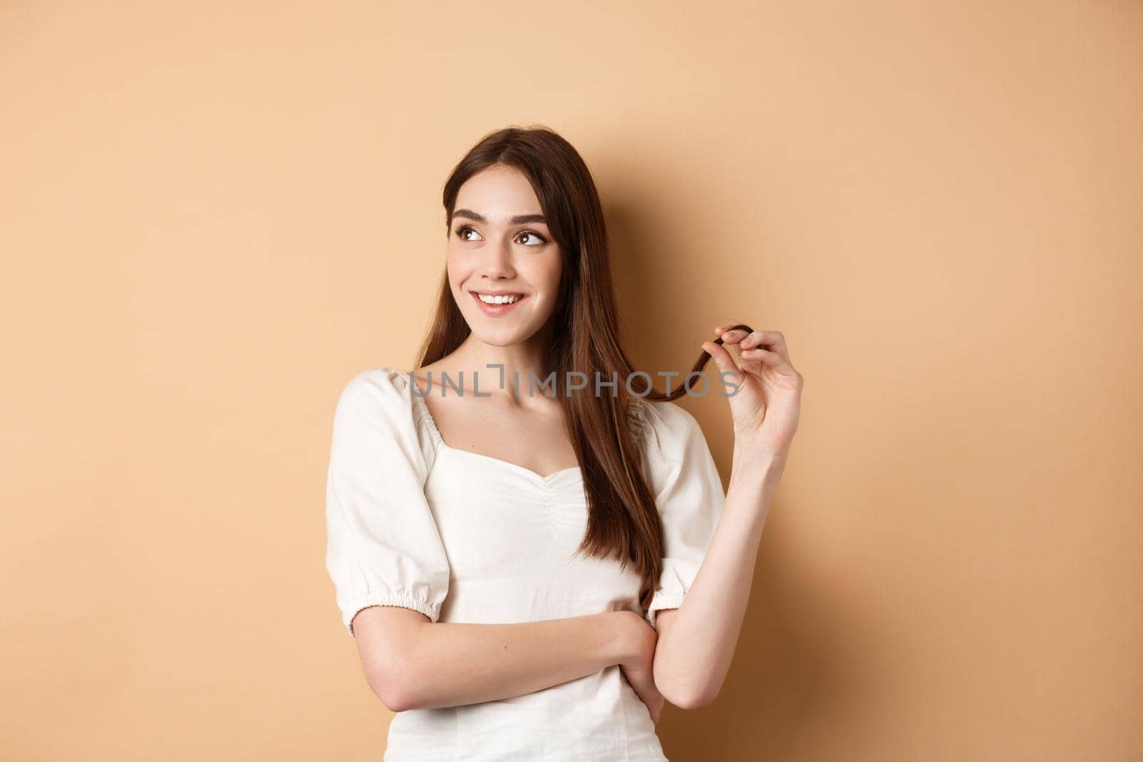 Romantic pensive girl thinking, looking left at empty space and playing with hair strand, smiling dreamy, standing in white dress on beige background.