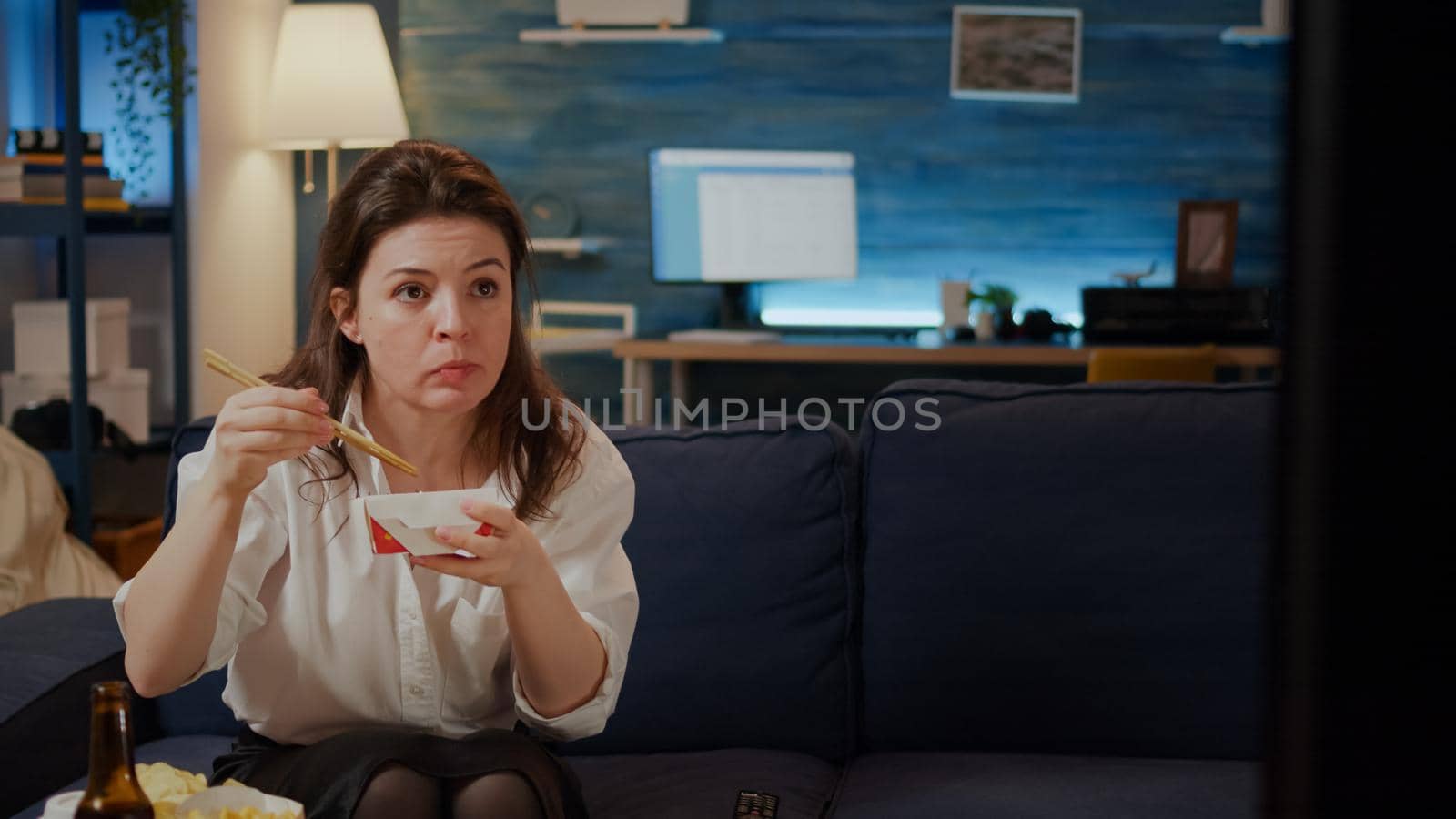 Person eating asian noodles with chopsticks on couch sitting in living room. Caucasian woman with chinese food box from delivery package enjoying meal while watching movie on TV screen