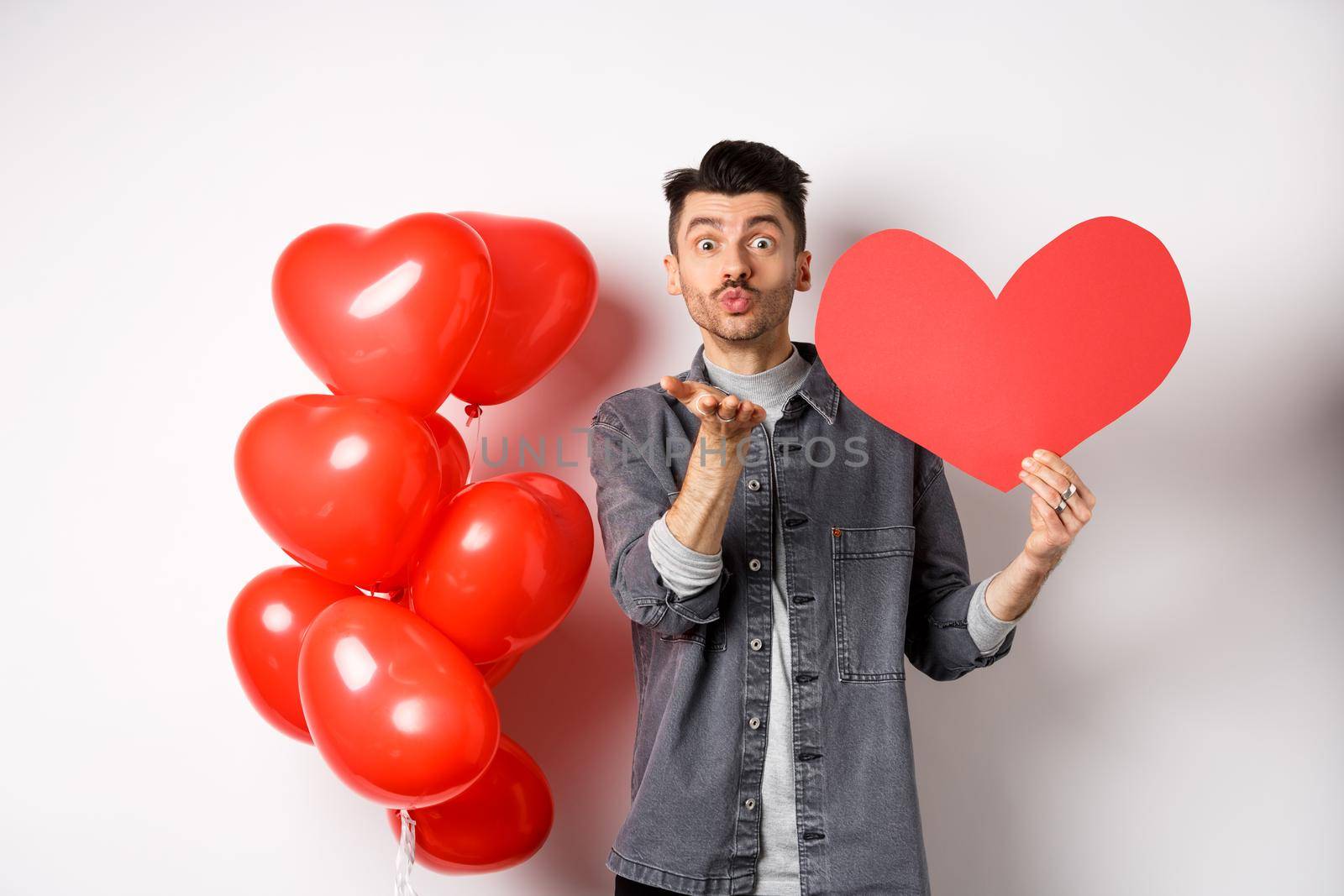 Valentines day and love concept. Romantic boy showing heart card and blowing air kiss at camera, looking with sympathy at lover, celebrating white day.