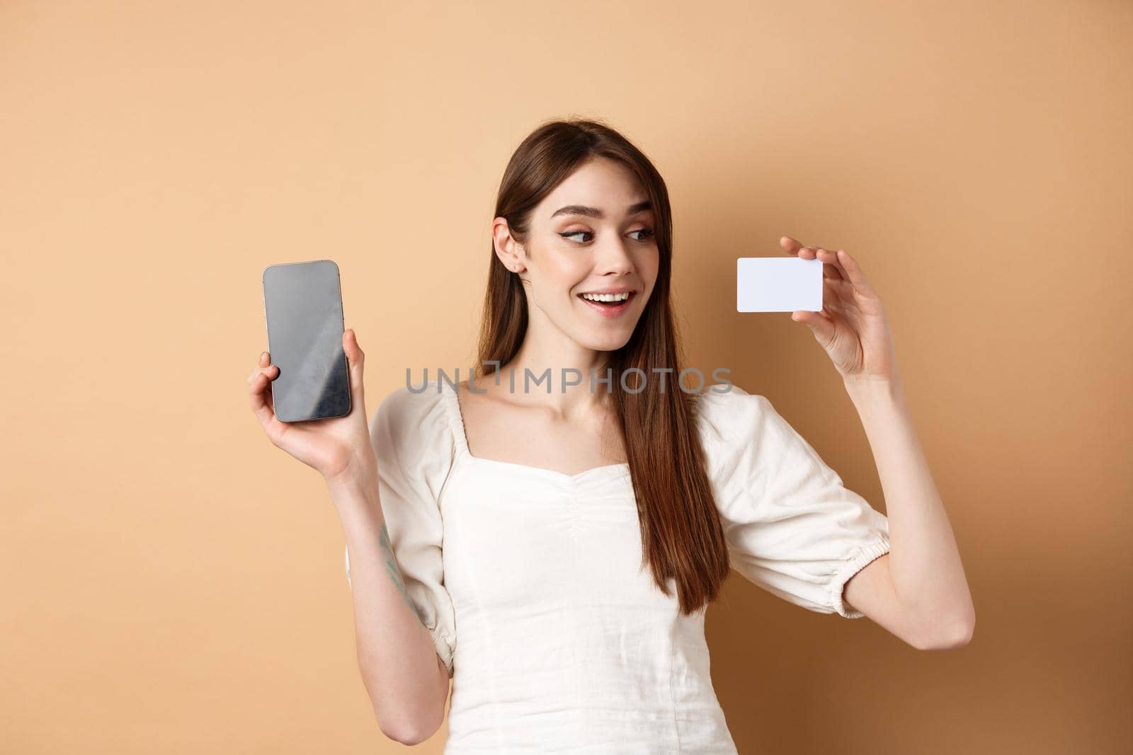 Young caucasian woman showing plastic credit card with pleased smile, demonstrate empty cellphone screen, standing on beige background by Benzoix