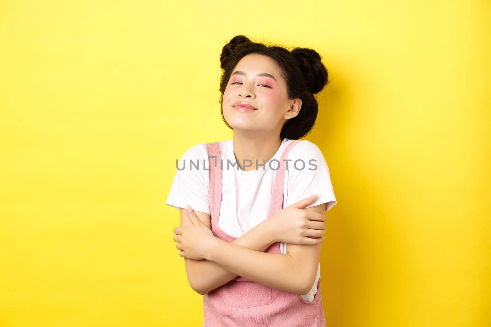 Summer lifestyle concept. Beautiful asian woman hugging herself and smiling romantic, embracing own body, standing with makeup on yellow background.