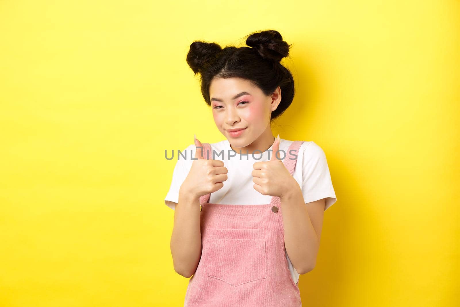 Cute teen asian girl with beauty makeup, showing thumbs up and smiling, praising and motivating you, making compliment, standing pleased on yellow background.